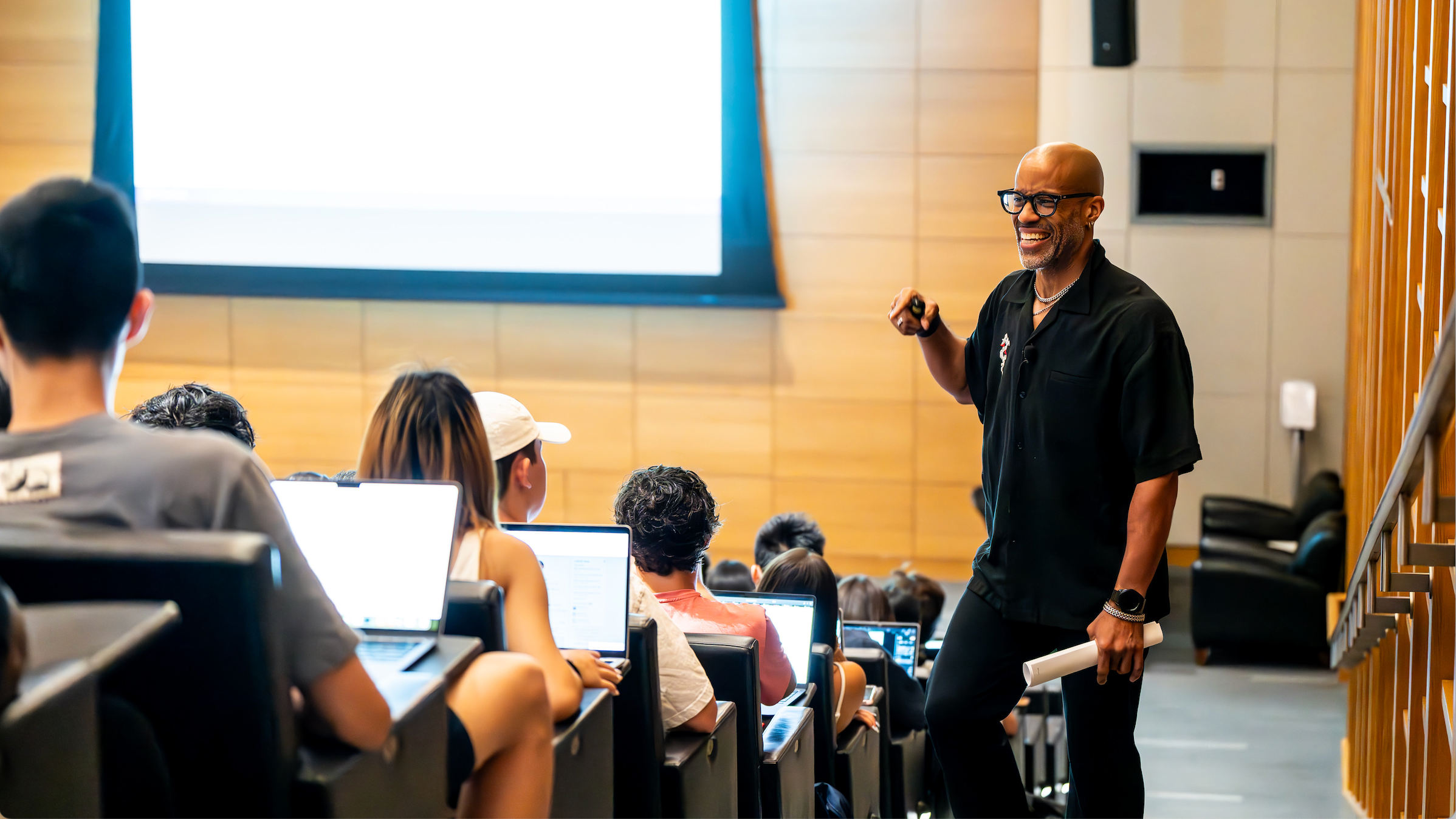 A smiling professor dressed in black talks to a lecture hall filled with high school students