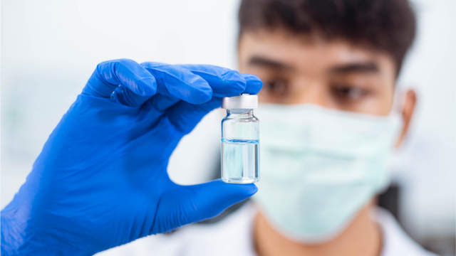 A person wearing a face mask and blue gloves is holding up a vial of clear liquid, possibly in a laboratory or medical setting.