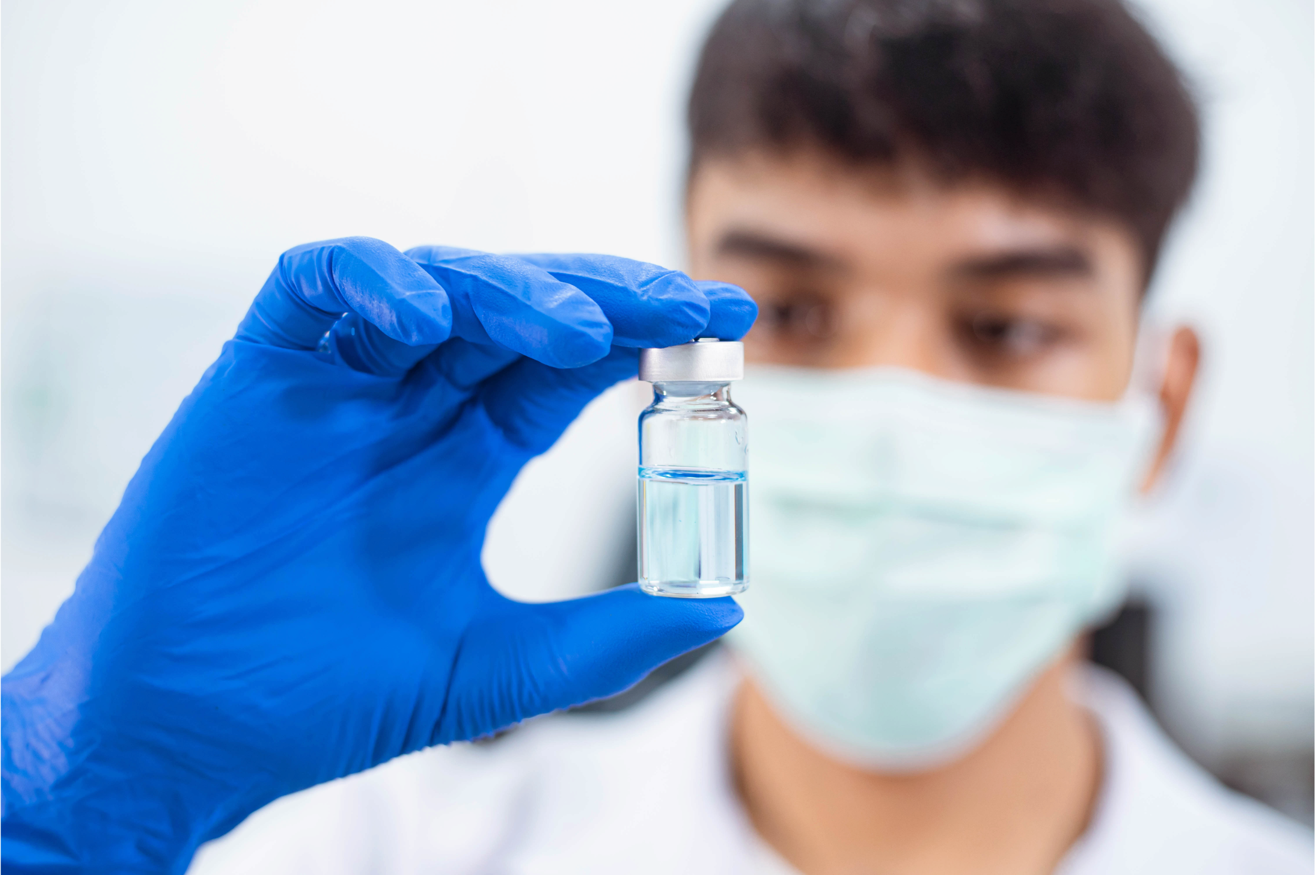 A person wearing a face mask and blue gloves is holding up a vial of clear liquid, possibly in a laboratory or medical setting.