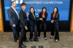 A group of people in business attire is standing in front of a display that reads "Global High School Investment Competition." They appear to be celebrating or posing for an achievement.