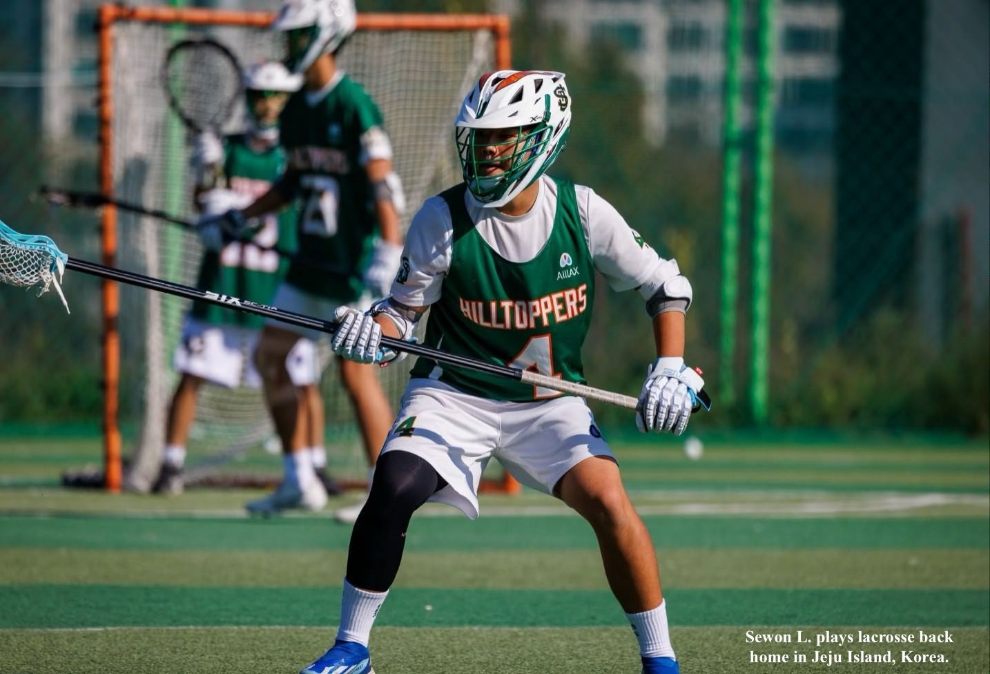 A sports player wearing a face mask and in white shirt, white shorts and green jersey holds a lacrosse stick.