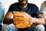 A person sitting holding a baseball glove, wearing a dark shirt and jeans, conveying a connection to baseball or sports.