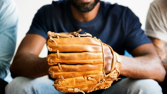 A person sitting holding a baseball glove, wearing a dark shirt and jeans, conveying a connection to baseball or sports.