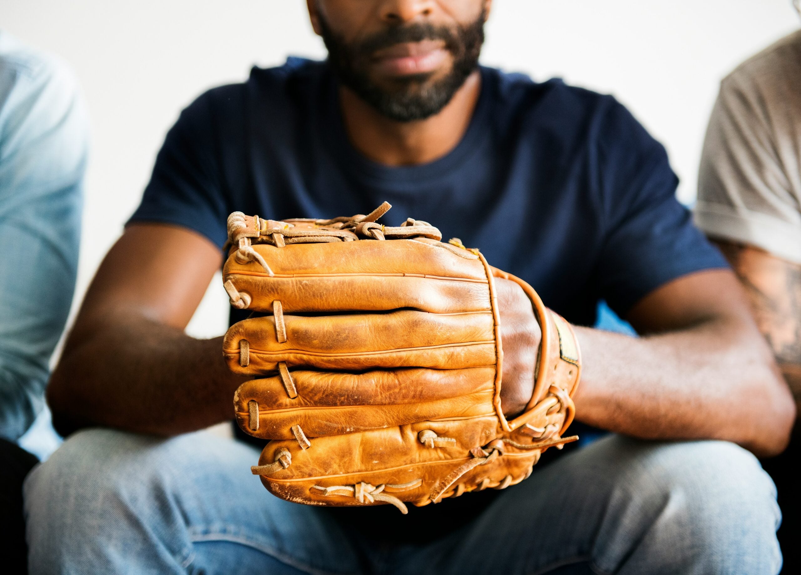 A person sitting holding a baseball glove, wearing a dark shirt and jeans, conveying a connection to baseball or sports.