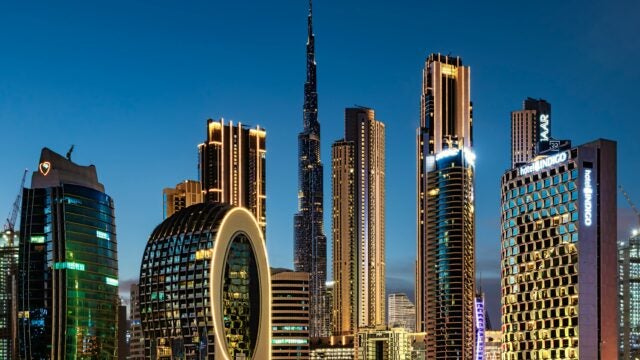 A city skyline at dusk featuring illuminated high-rise buildings, including a distinctive skyscraper in the center.