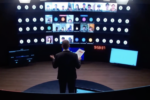 A person stands in front of a large curved display showing multiple video call participants in a virtual meeting. The setting resembles a modern conference or presentation room.