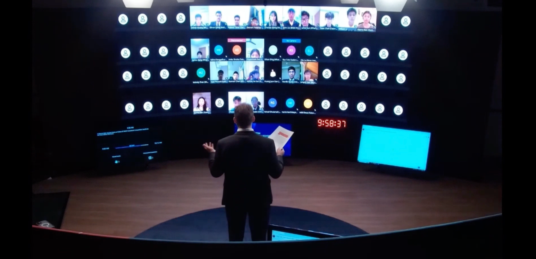 A person stands in front of a large curved display showing multiple video call participants in a virtual meeting. The setting resembles a modern conference or presentation room.