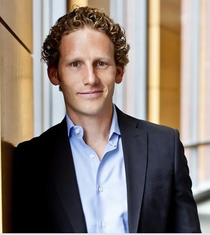 man in light blue button-down shirt and dark blue blazer leaning against a wall and smiling.