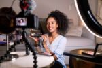 A person sitting in front of a camera setup and ring light, demonstrating makeup application with a palette and brush, suggesting a beauty tutorial video.