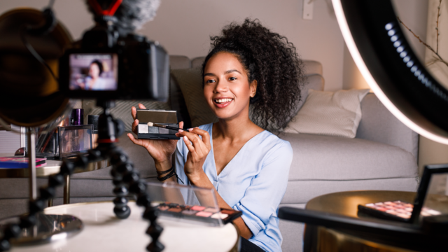 A person sitting in front of a camera setup and ring light, demonstrating makeup application with a palette and brush, suggesting a beauty tutorial video.