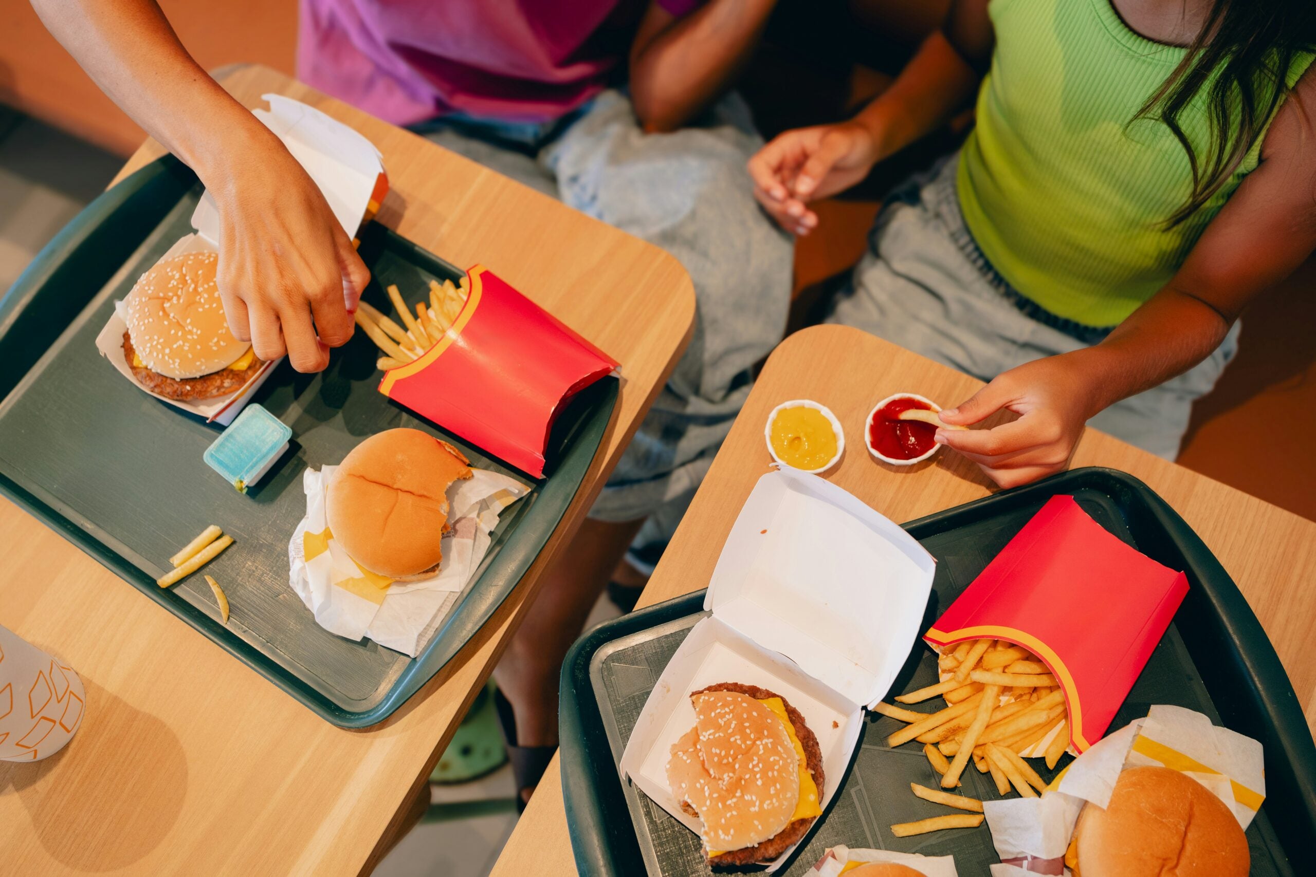 Two people sitting at a table eating fast food, featuring burgers and fries with ketchup and mustard sauces on the side.