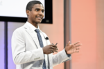 young man wearing an off-white blazer, white shirt and blue tie standing on a stage speaking with his hands raised.