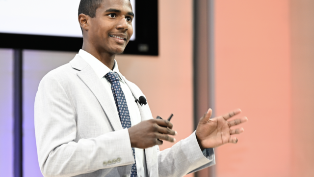young man wearing an off-white blazer, white shirt and blue tie standing on a stage speaking with his hands raised.