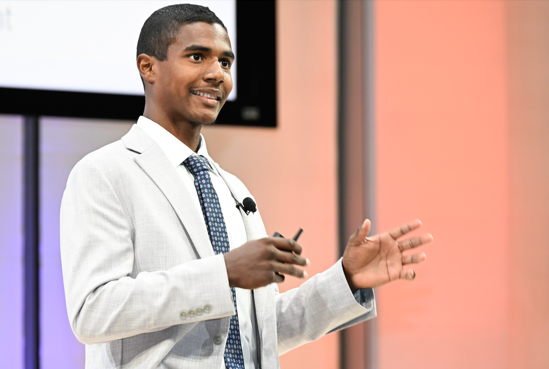 young man wearing an off-white blazer, white shirt and blue tie standing on a stage speaking with his hands raised.