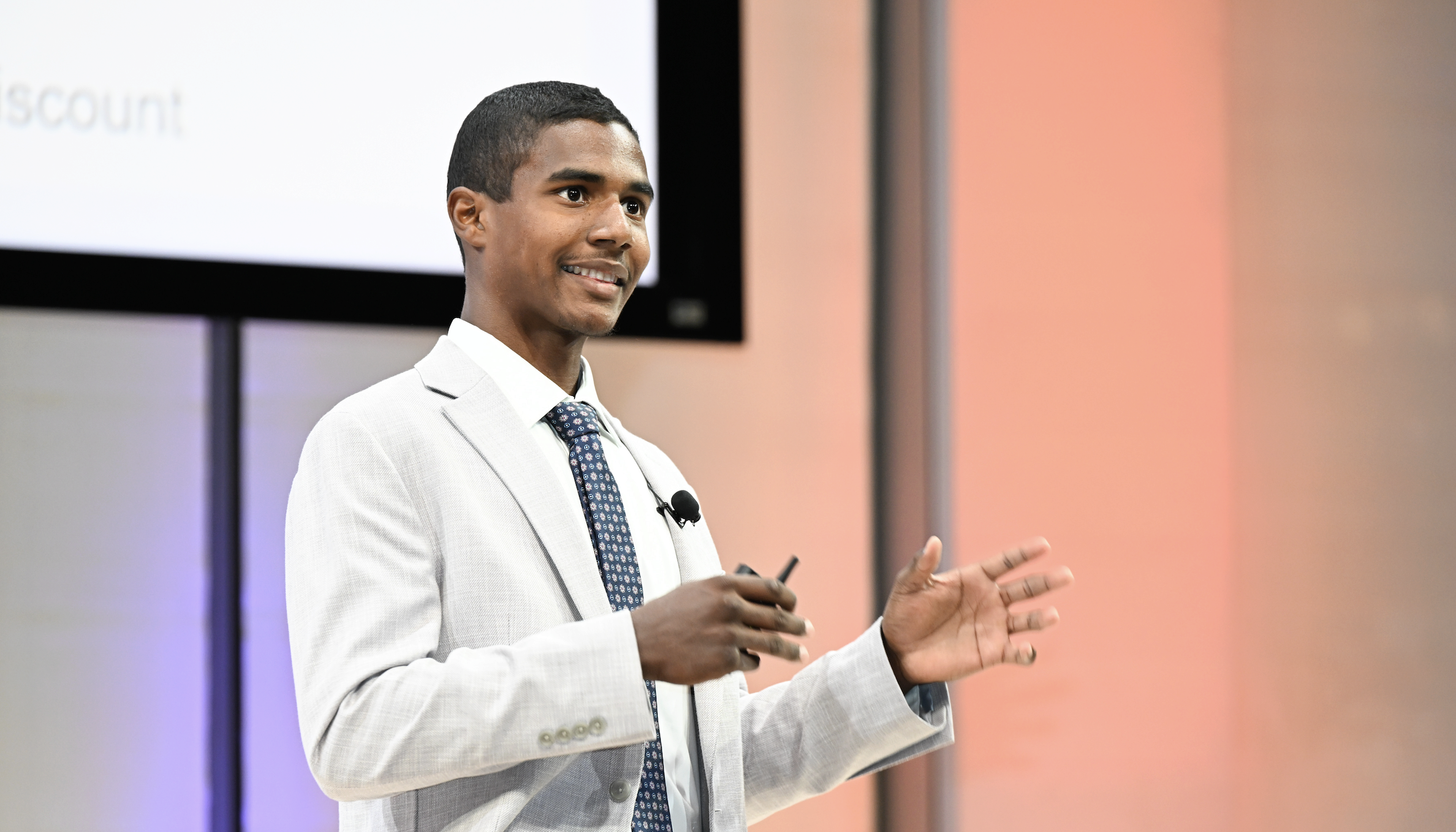 A person in a light gray suit and polka dot tie is speaking and gesturing with hands, equipped with a microphone headset, during a presentation.