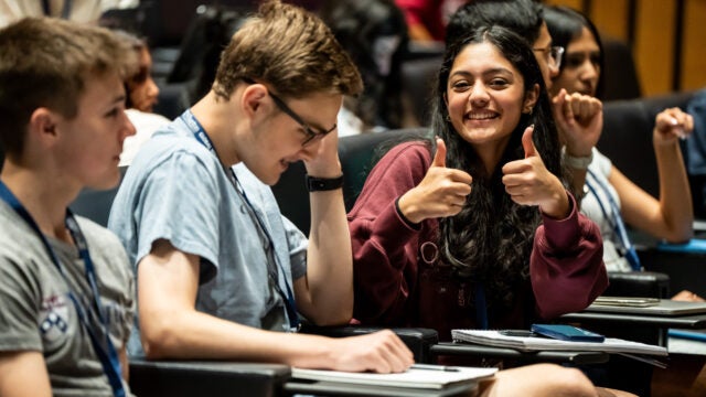 A small group of young adults is seated in what appears to be a lecture hall. One individual is smiling broadly and giving a thumbs-up, suggesting a positive and engaged atmosphere.