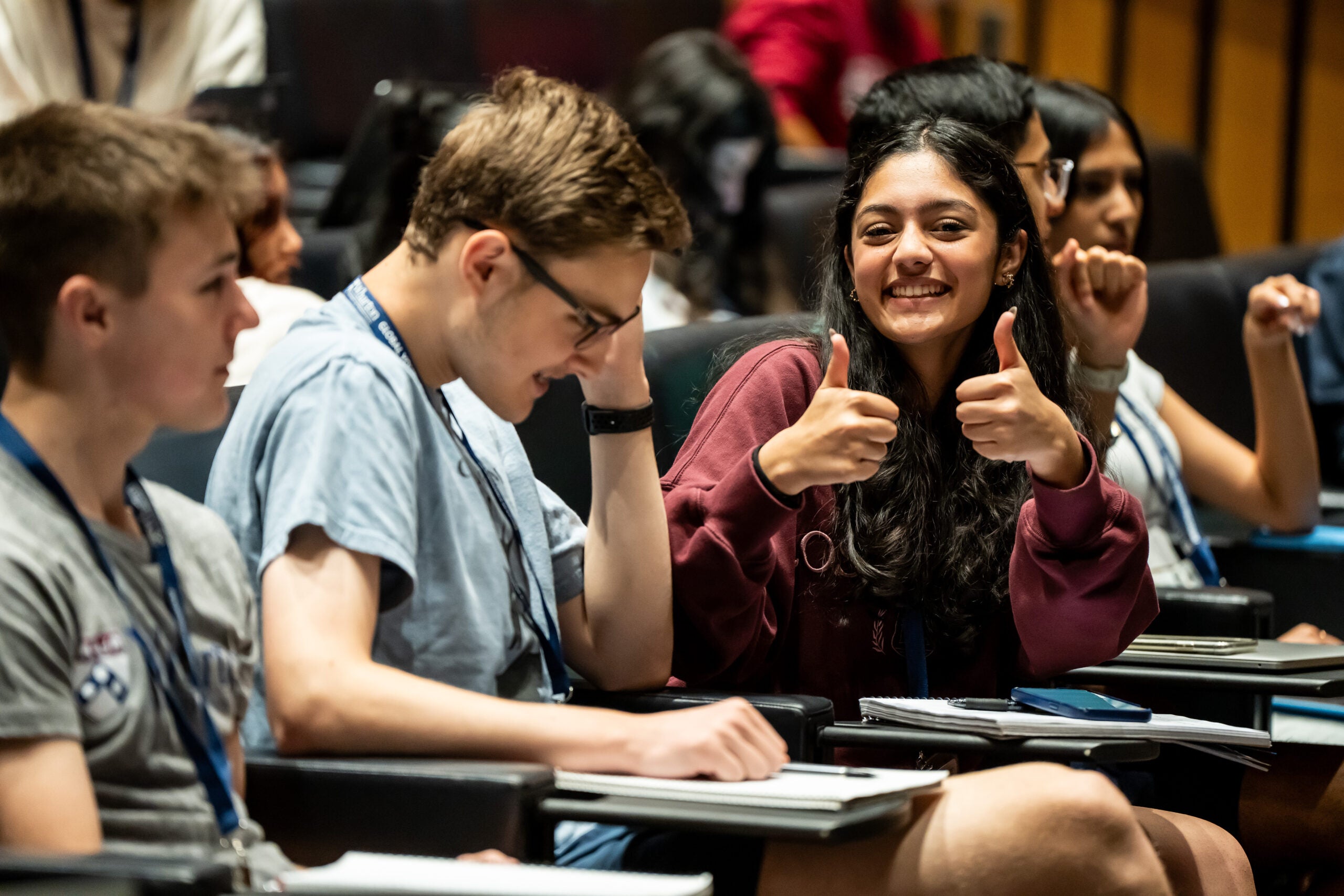 A small group of young adults is seated in what appears to be a lecture hall. One individual is smiling broadly and giving a thumbs-up, suggesting a positive and engaged atmosphere.