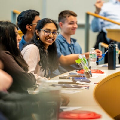 250425 Wgyp Invest 1250 A group of students sitting in a lecture hall, smiling and engaged. Tables are equipped with water bottles, papers, and small items.