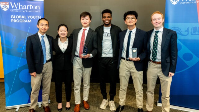 A group of six people standing in a line, dressed in formal and business-casual attire. Two individuals hold awards. Blue Wharton Global Youth Program banners are in the background.