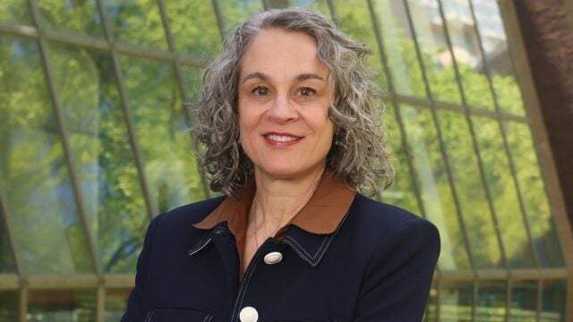 Headshot of a person with curly gray hair, wearing a dark jacket with a brown collar. The background features large windows with a view of greenery.