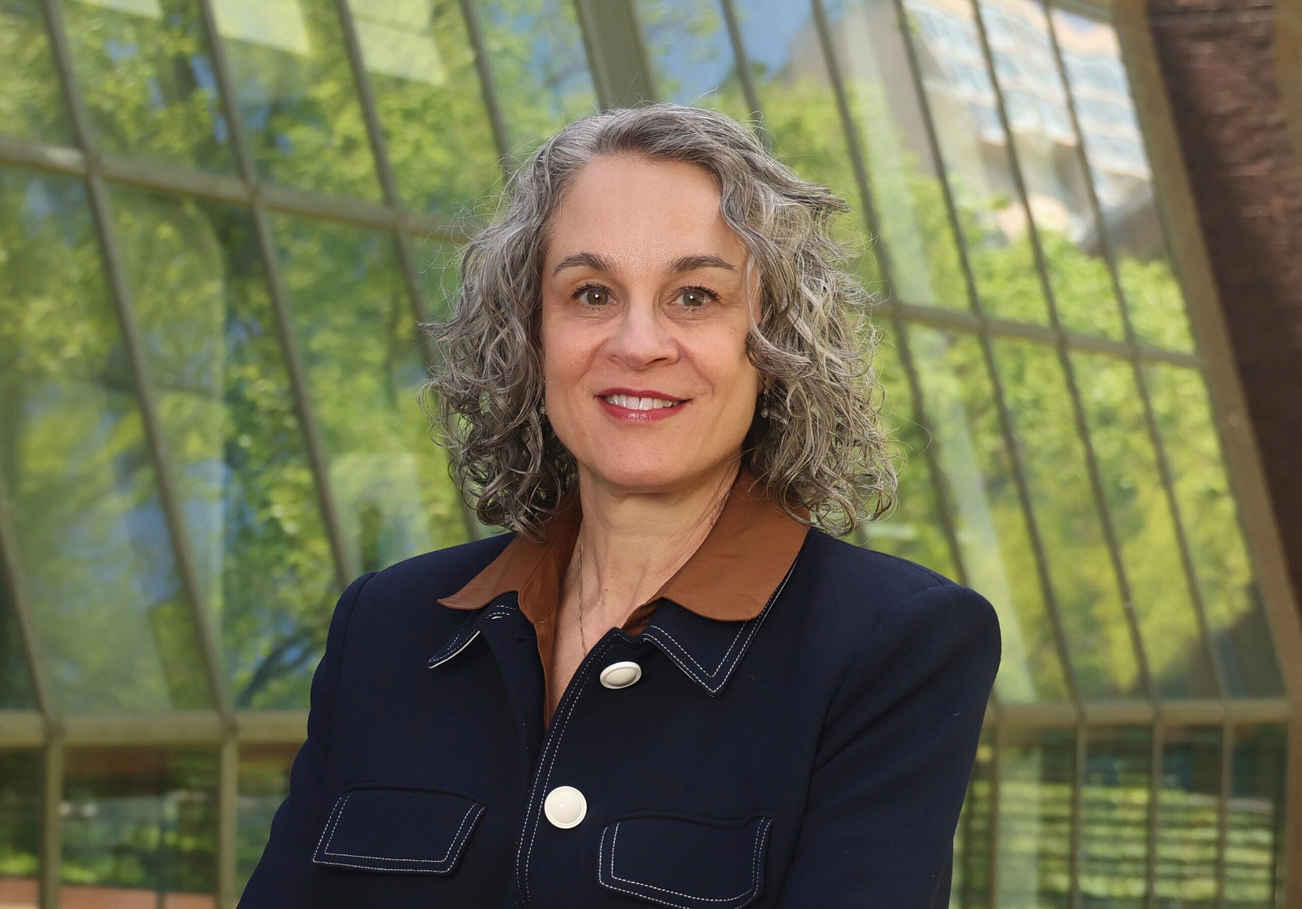 Headshot of a person with curly gray hair, wearing a dark jacket with a brown collar. The background features large windows with a view of greenery.