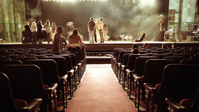 An auditorium with a red carpet aisle leading to a stage. Several people are gathered on the stage under bright stage lights, while others sit in the audience seats.