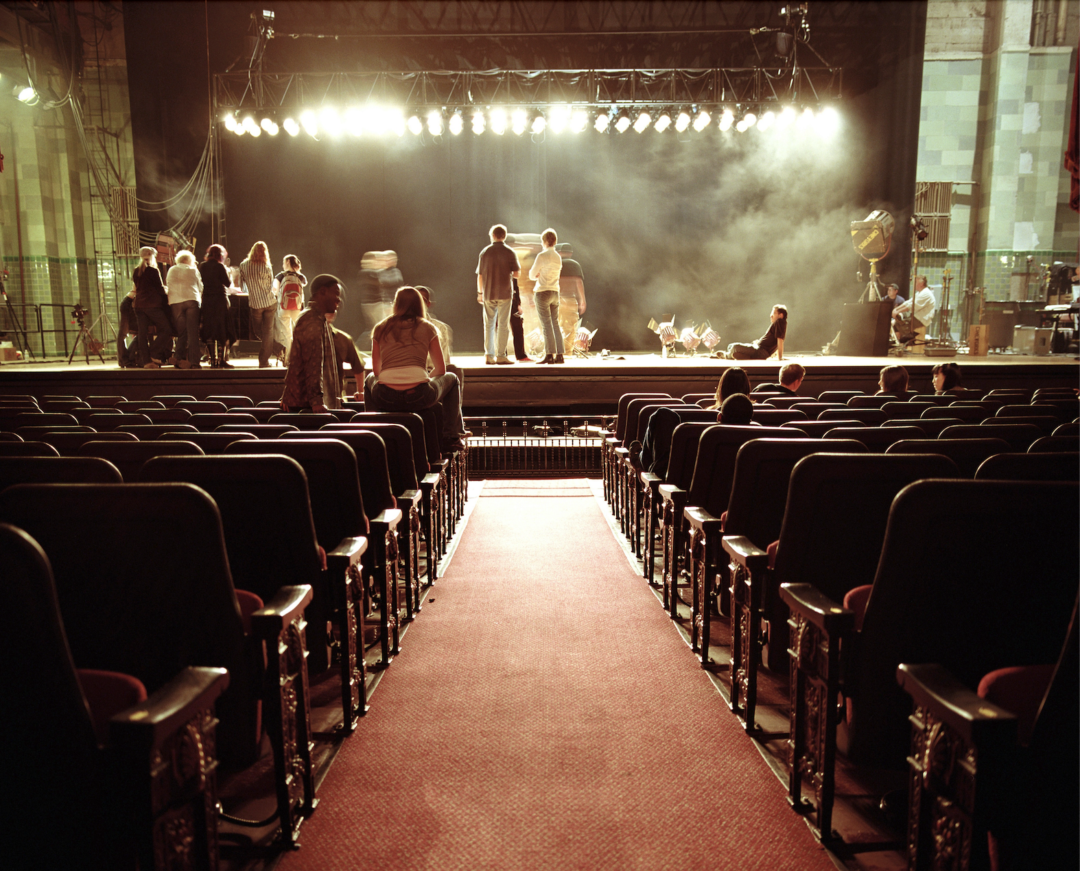 An auditorium with a red carpet aisle leading to a stage. Several people are gathered on the stage under bright stage lights, while others sit in the audience seats.