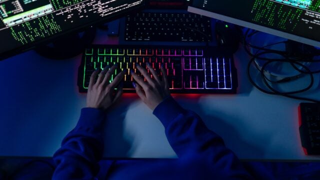 A person typing on a colorful backlit keyboard, surrounded by multiple monitors displaying code or data. This represents a programmer or hacker at work.