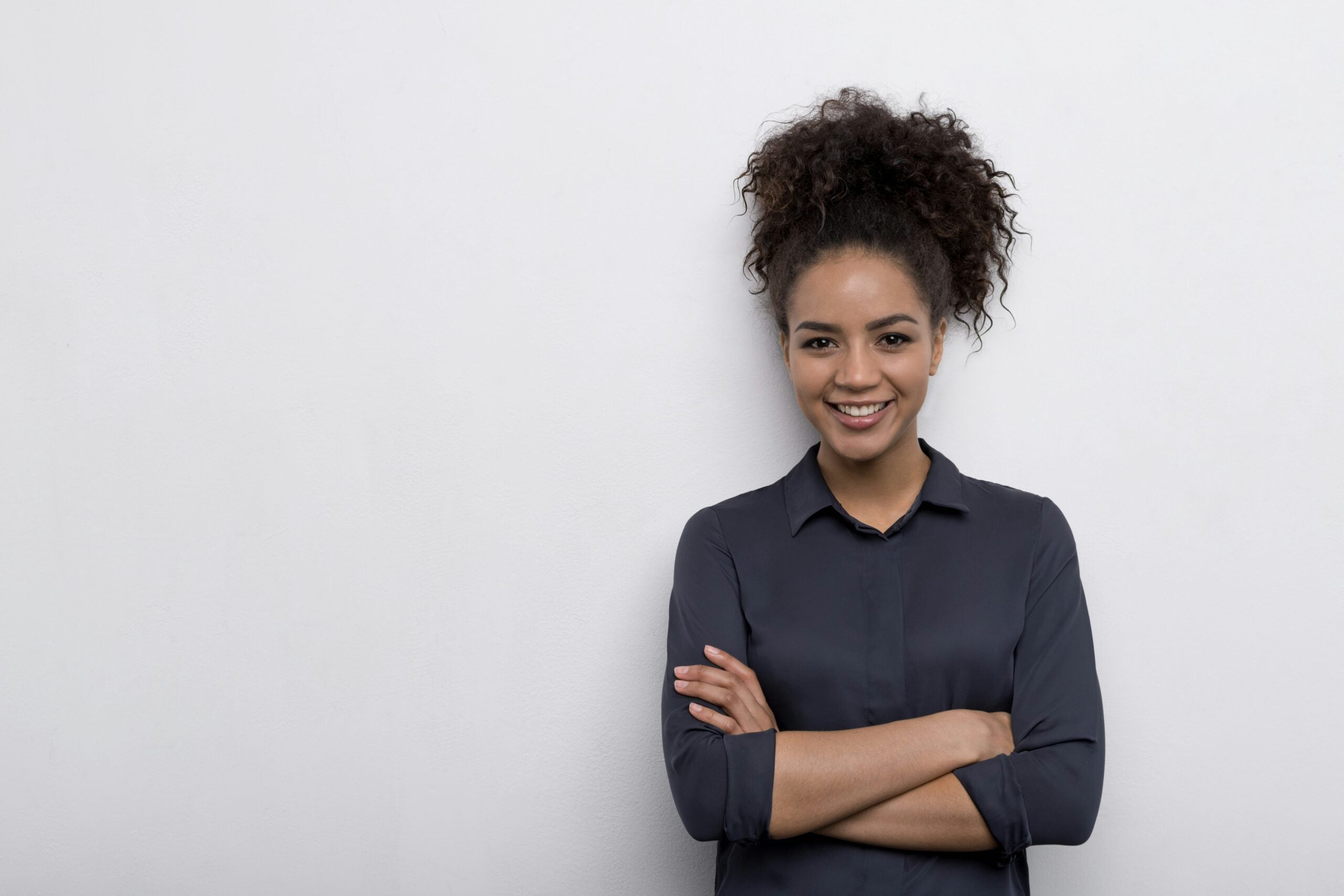 A person with dark, curly hair tied up, smiling with arms crossed, wearing a dark long-sleeve shirt, standing against a plain light background.
