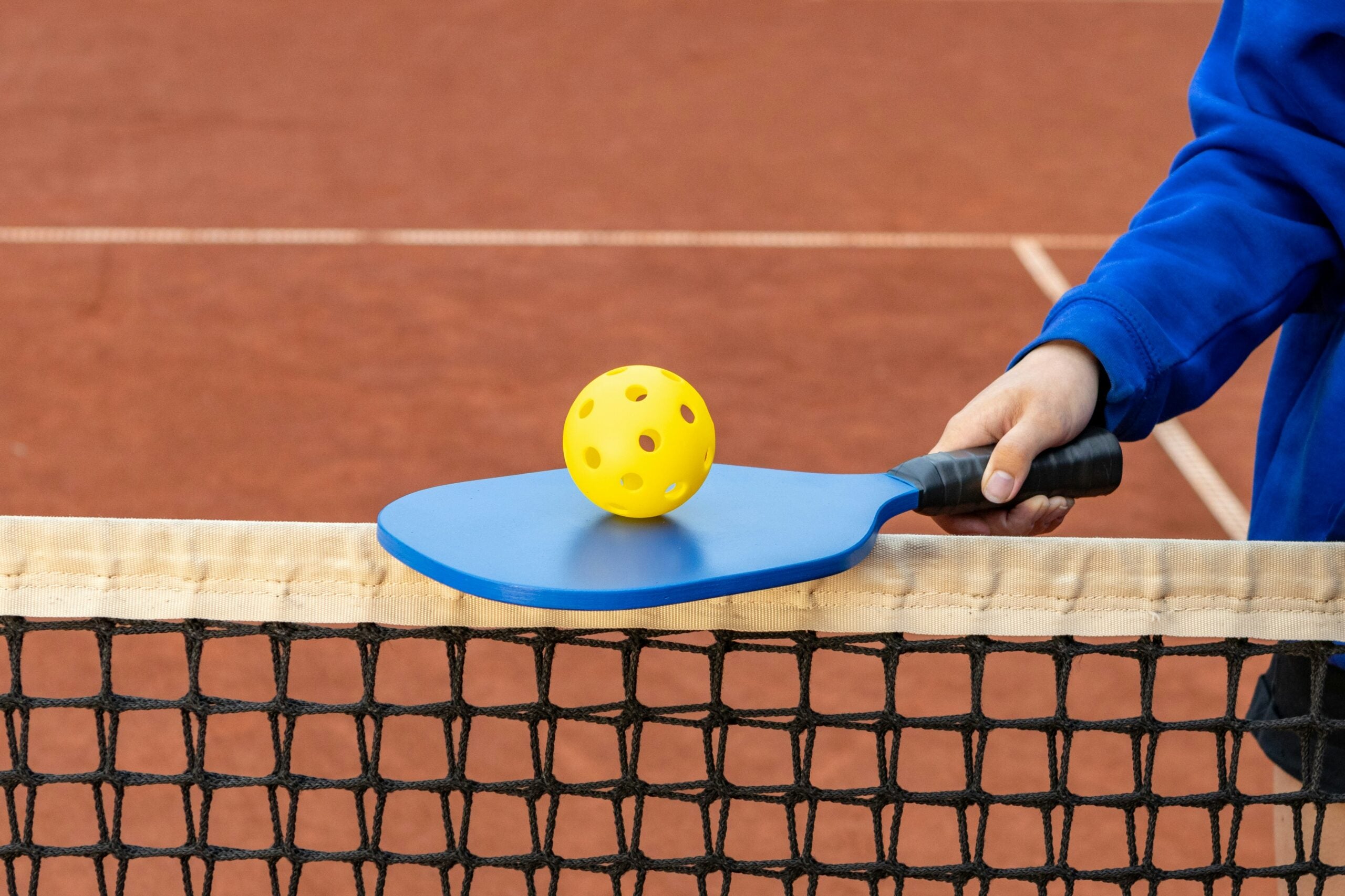 A person holding a blue pickleball paddle with a yellow perforated ball resting on it, above a net on a clay court.