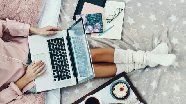 A person sitting on a bed with a star-patterned blanket, using a laptop. Nearby are a tablet, notebooks, glasses, and a pen. A tray holds a cupcake, coffee, and a small bundle of lavender. The scene suggests a cozy