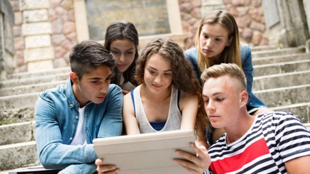 A small group of young people sitting on outdoor steps, closely gathered around a tablet, engaged in what they are viewing together.
