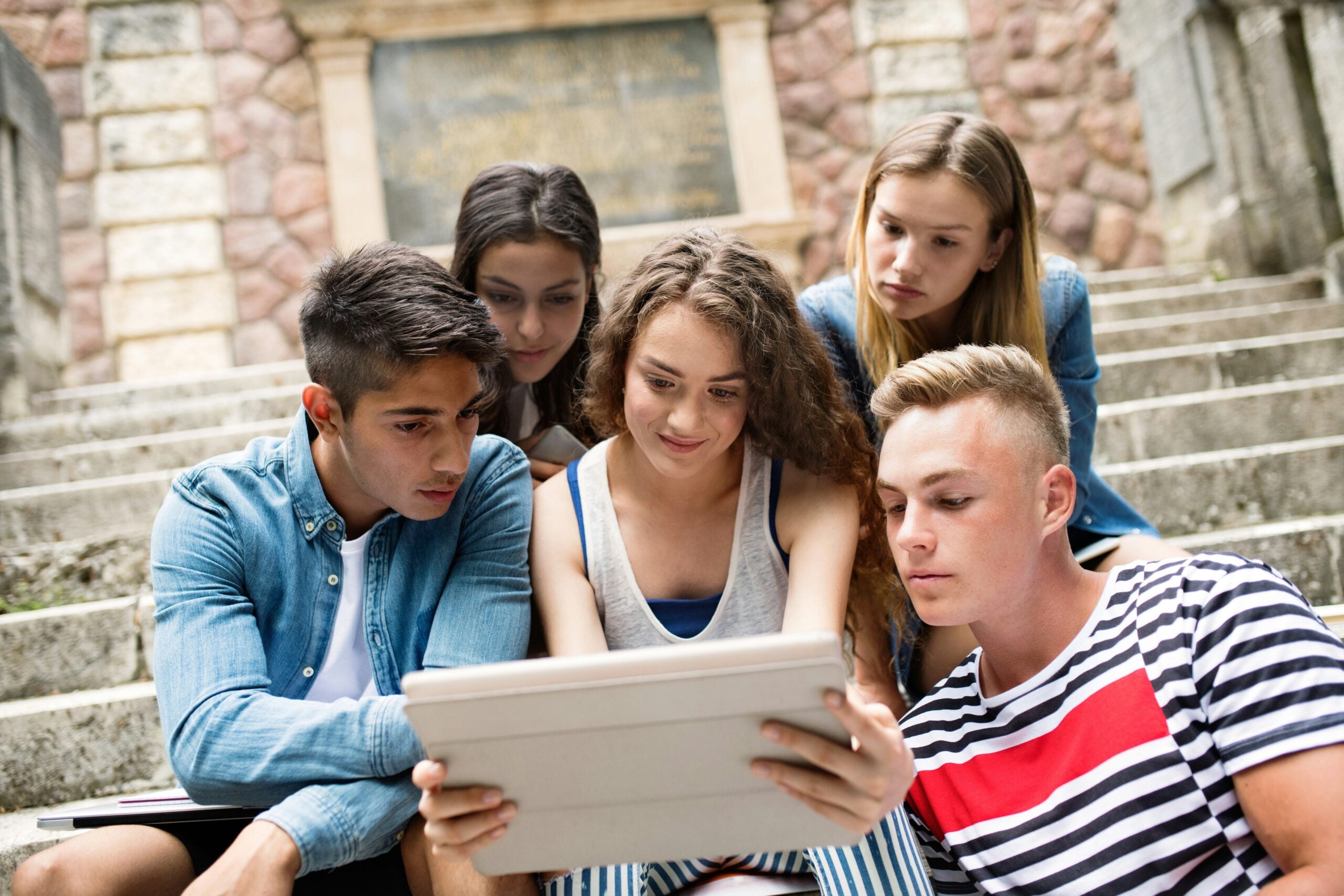 A small group of young people sitting on outdoor steps, closely gathered around a tablet, engaged in what they are viewing together.