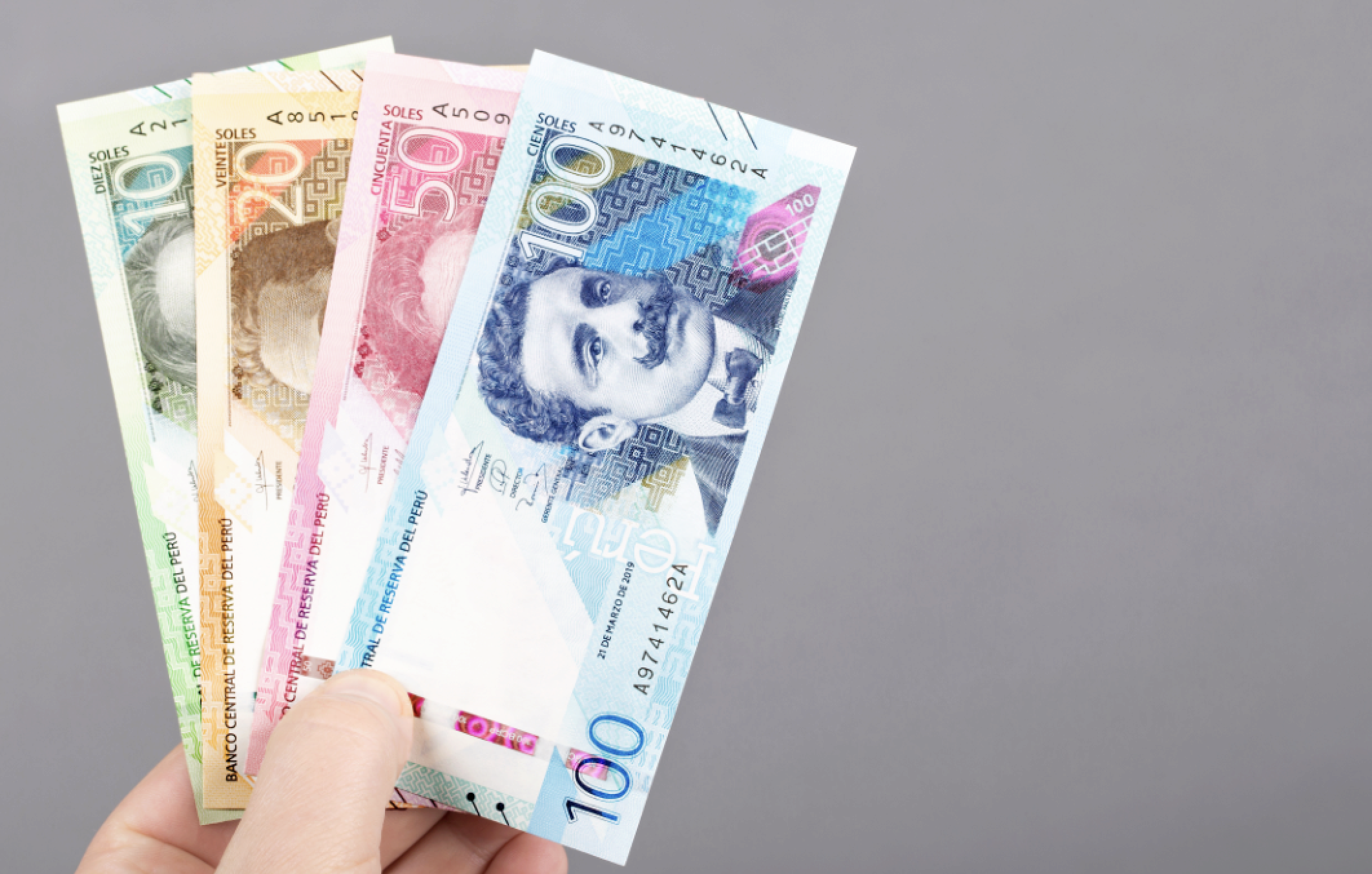 A hand holding a variety of Peruvian banknotes, including 10, 20, 50, and 100 Soles, on a plain background.