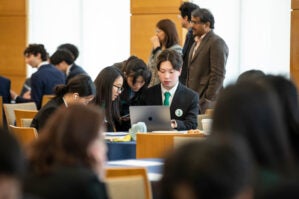 230422 WGYP Investment 0002 A group of young adults in formal attire collaborates around a laptop in a seminar or conference setting, with others in the background.