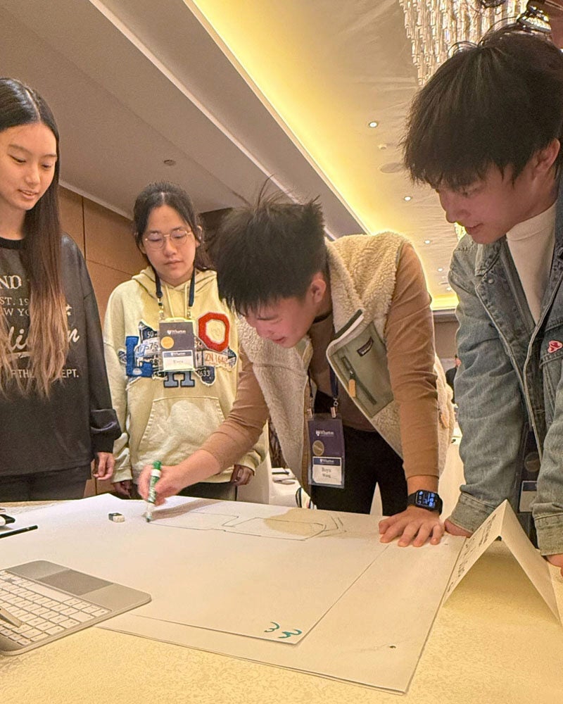 A small group of people gather around a table, engaging in a collaborative activity. One person is drawing on a large sheet of paper.