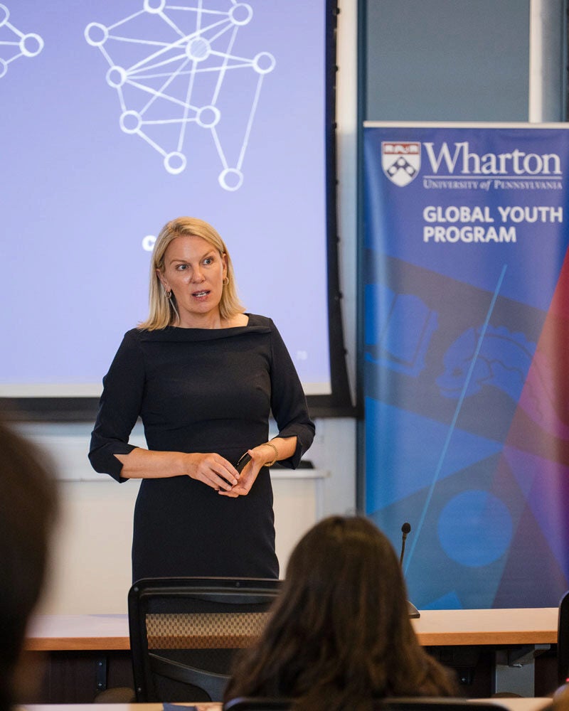 A person giving a presentation in a classroom, standing in front of a screen with a network diagram. A sign reads "Wharton Global Youth Program."