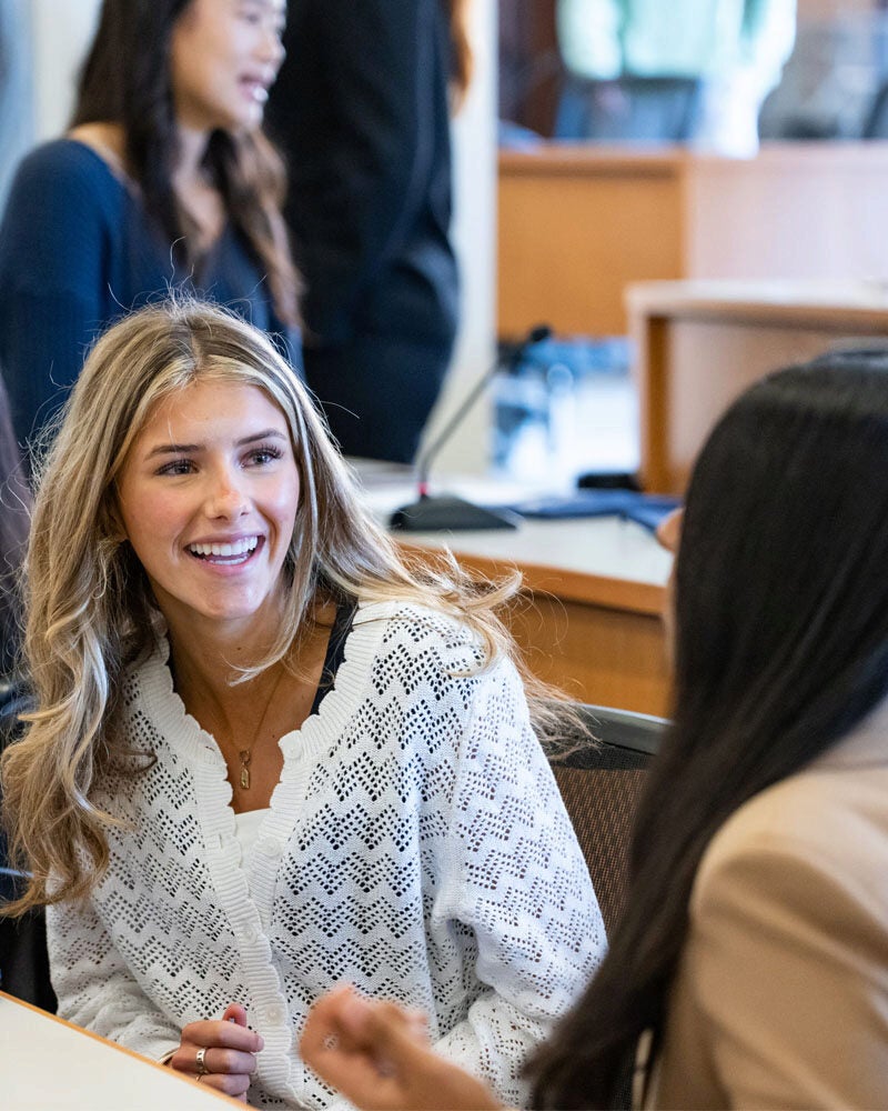 A few people in a professional or educational setting, with one person in the foreground smiling and engaging in conversation.