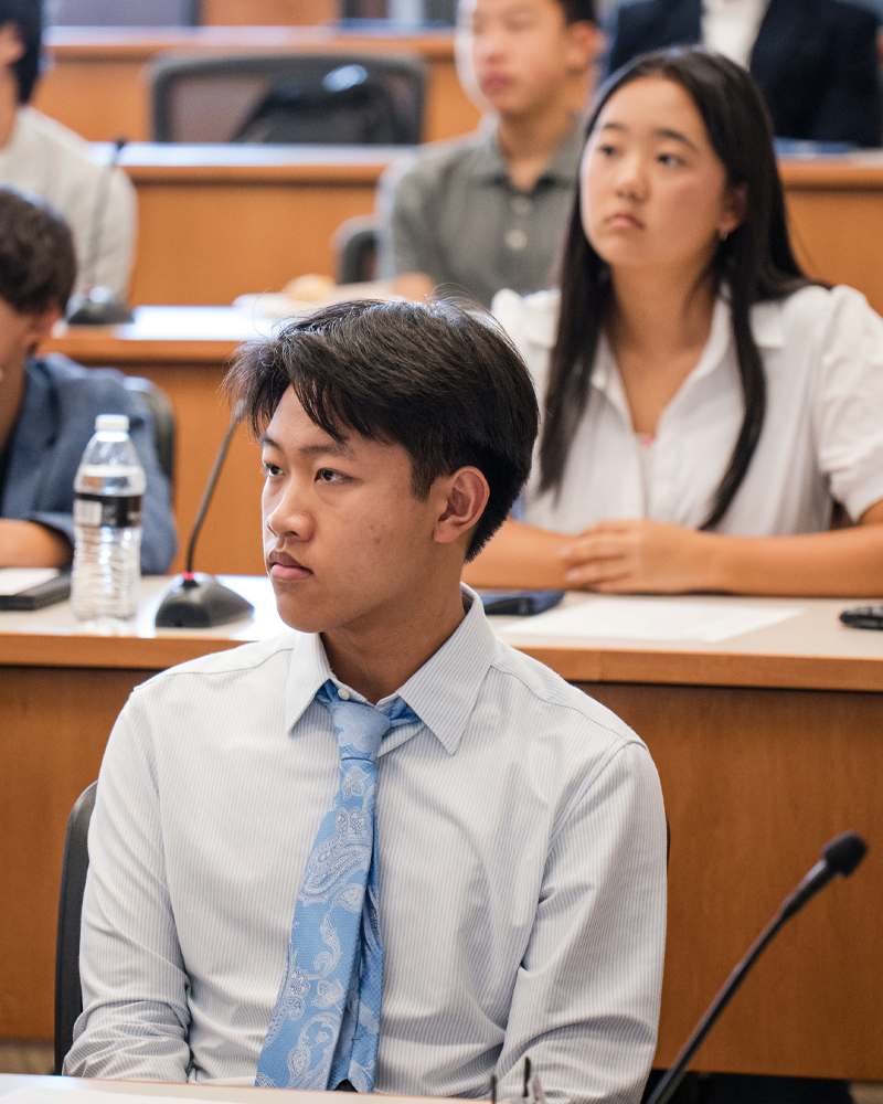 A few students sitting in a classroom, attentively listening, with one person in the foreground wearing a tie and a blue shirt.