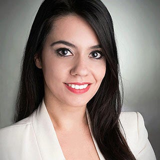 Headshot of a person with long dark hair, wearing a light-colored blazer and smiling against a neutral background.